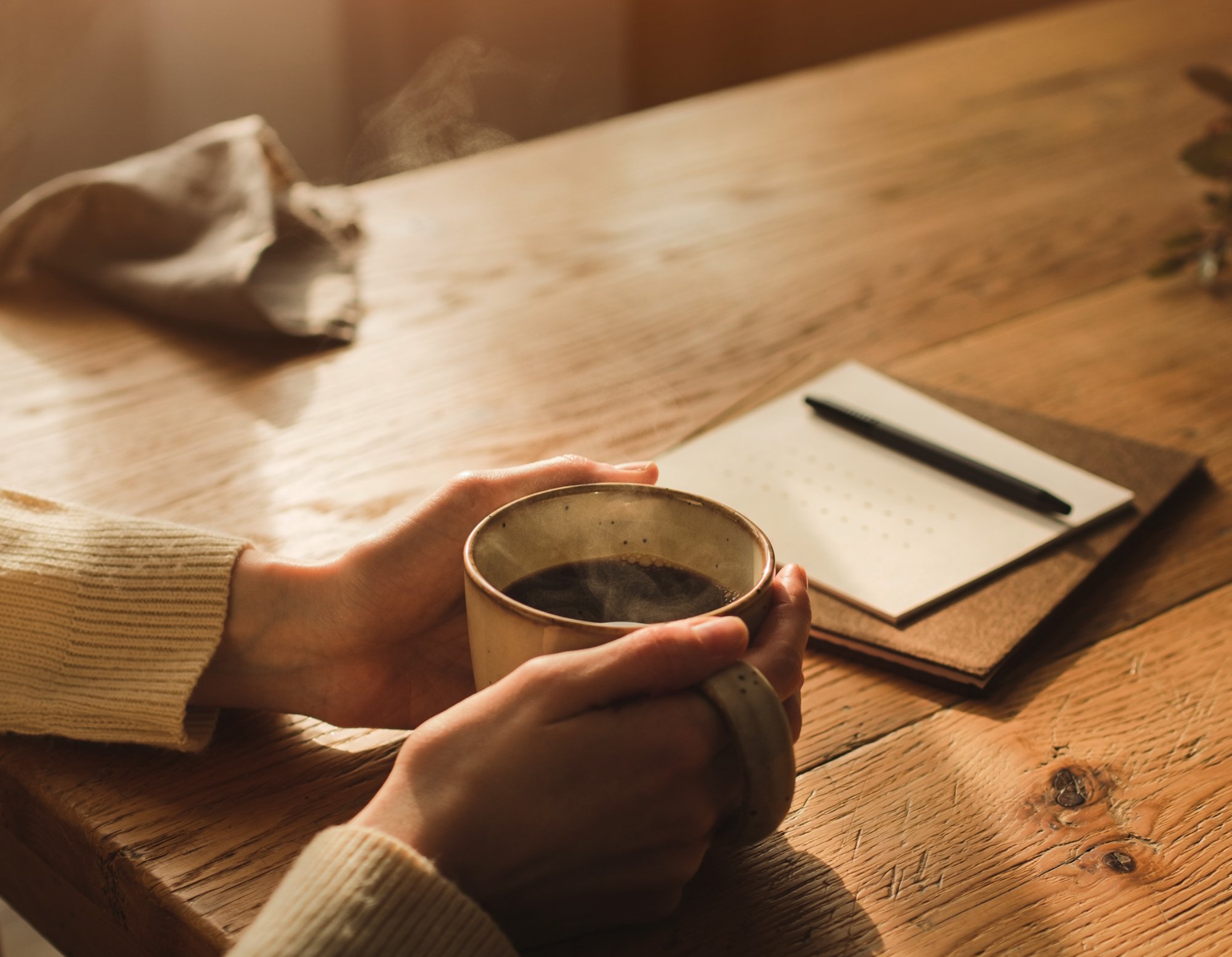 Hands holding a warm cup of coffee beside a notebook